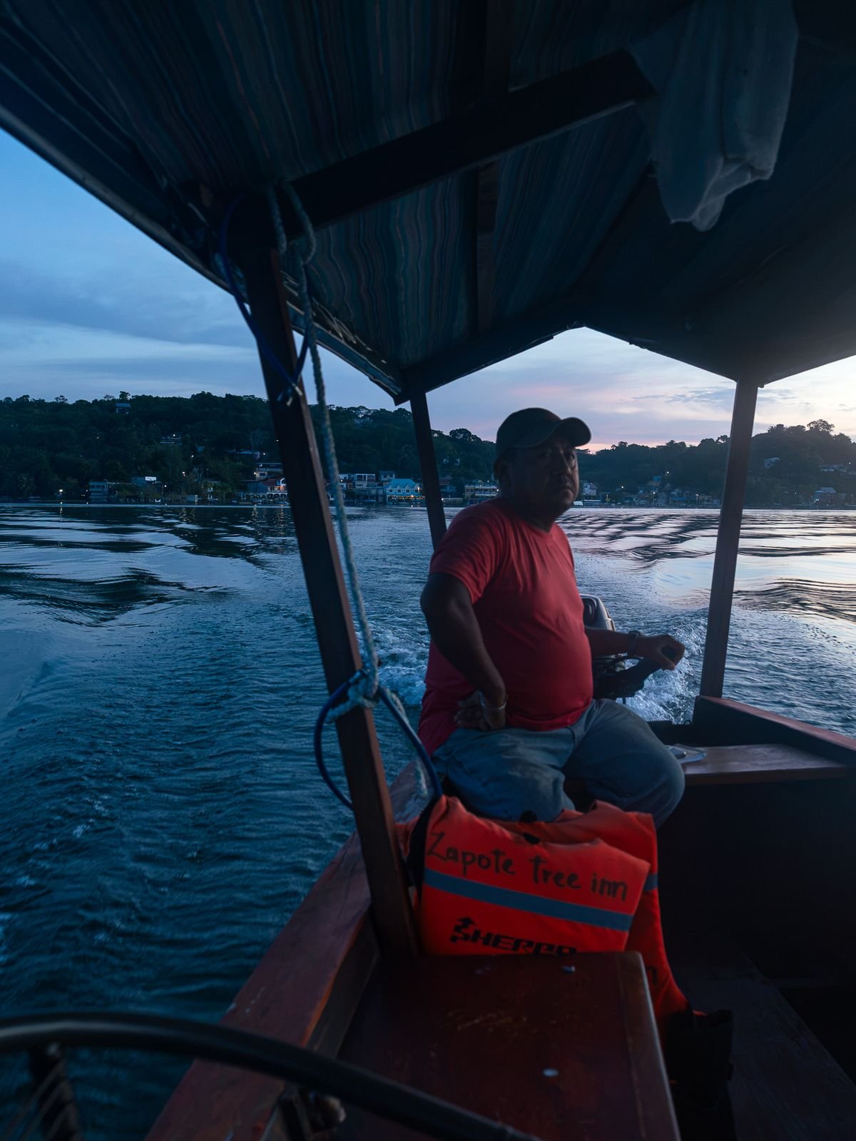 A boat driver in a red shirt steers a covered boat across a lake at dusk.