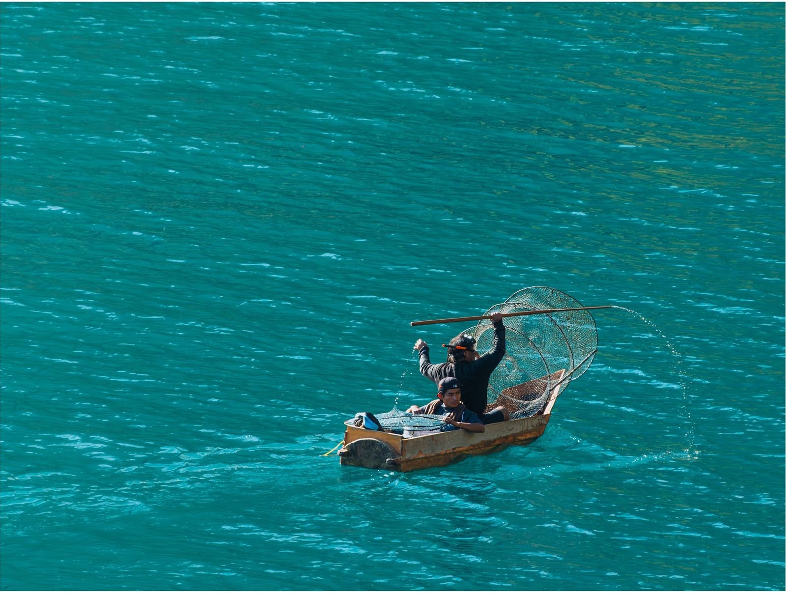 A fisherman in a small wooden boat throws a net over bright turquoise water.