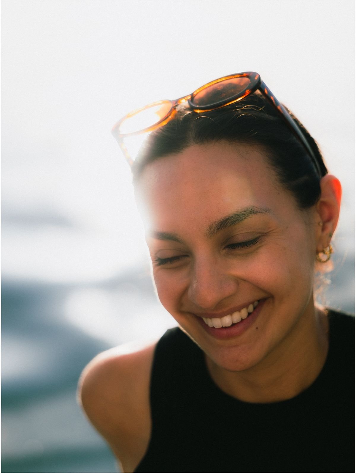 A woman with sunglasses pushed up on her head laughs and looks down, against soft, bright, blurred water.
