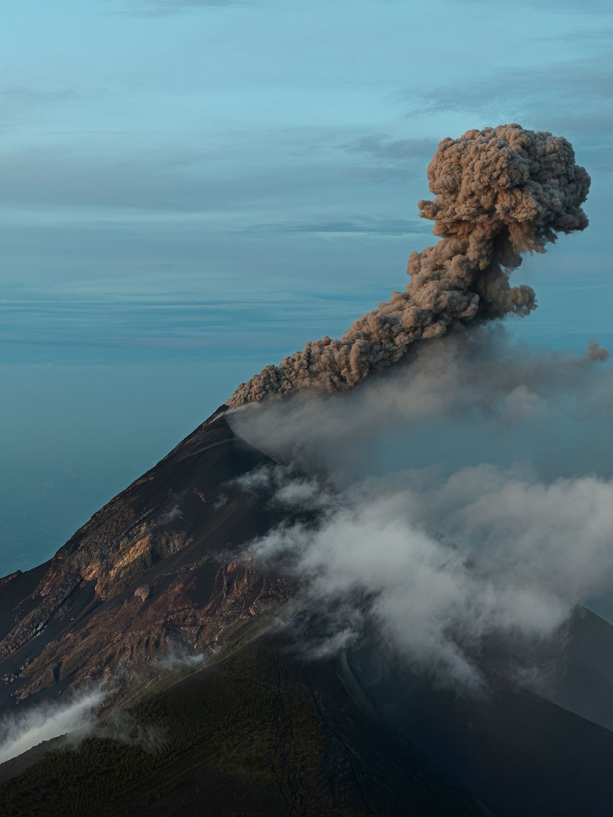 A volcano sends a tall column of gray ash into a clear blue sky while clouds drift across its slope.