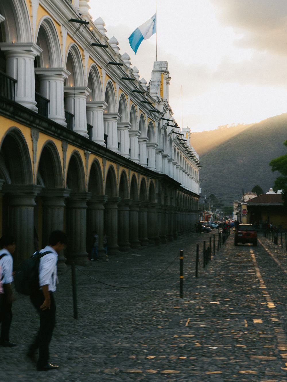 Students in white shirts walk along a cobblestone street beside a yellow colonial building with arches, under the Guatemalan flag.