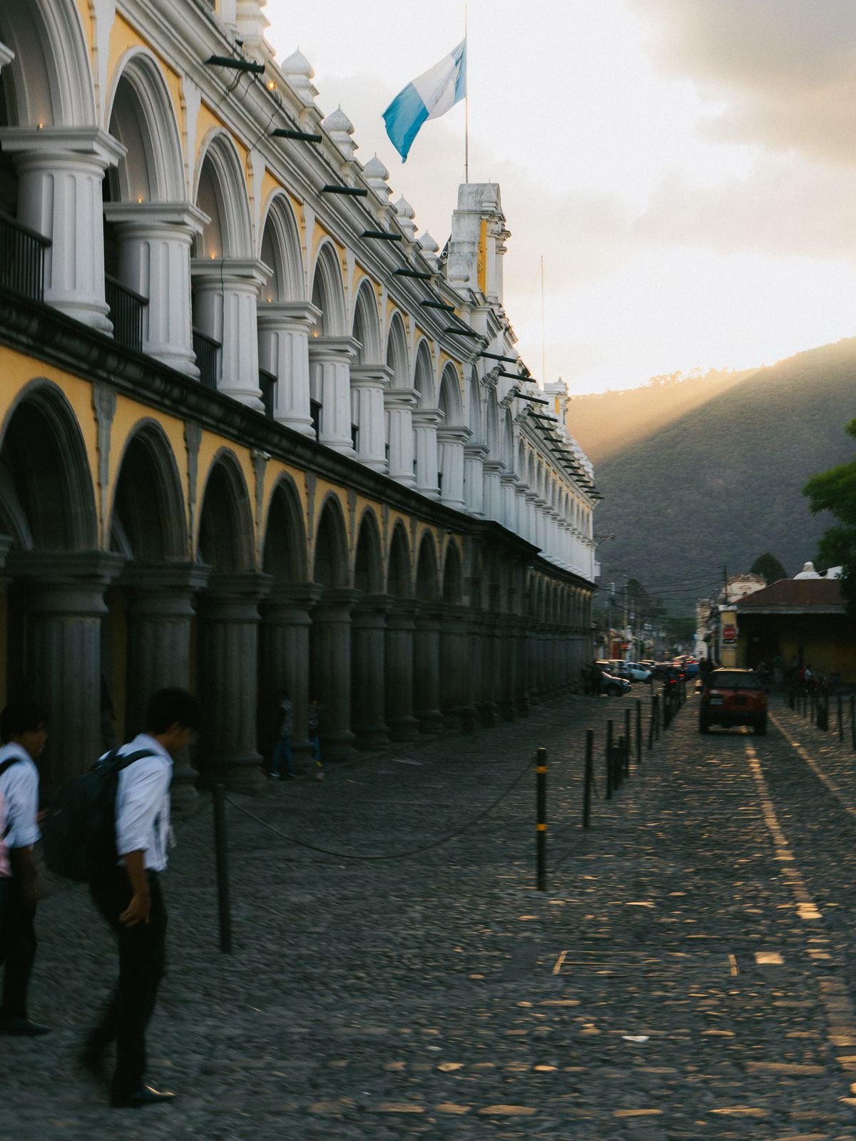 Students in white shirts walk along a cobblestone street beside a yellow colonial building with arches, under the Guatemalan flag.