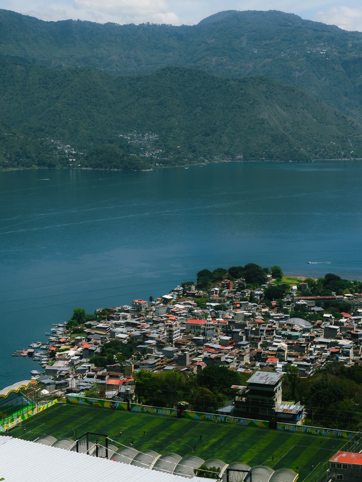 A town on the shore of a large lake, seen from above, with a soccer field in front and green mountains across the water.
