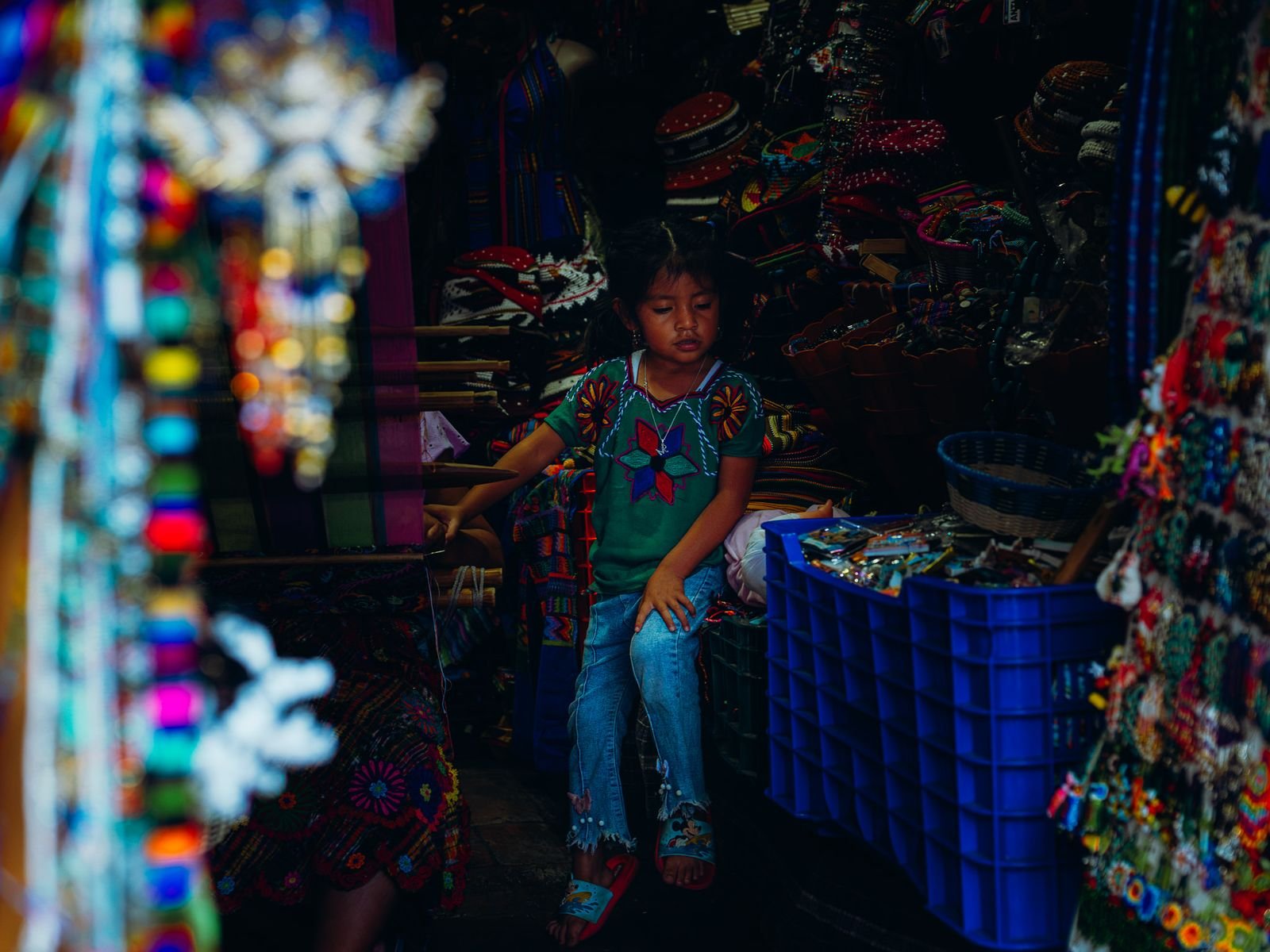 A girl sits in a market stall surrounded by colorful woven goods and beaded souvenirs.
