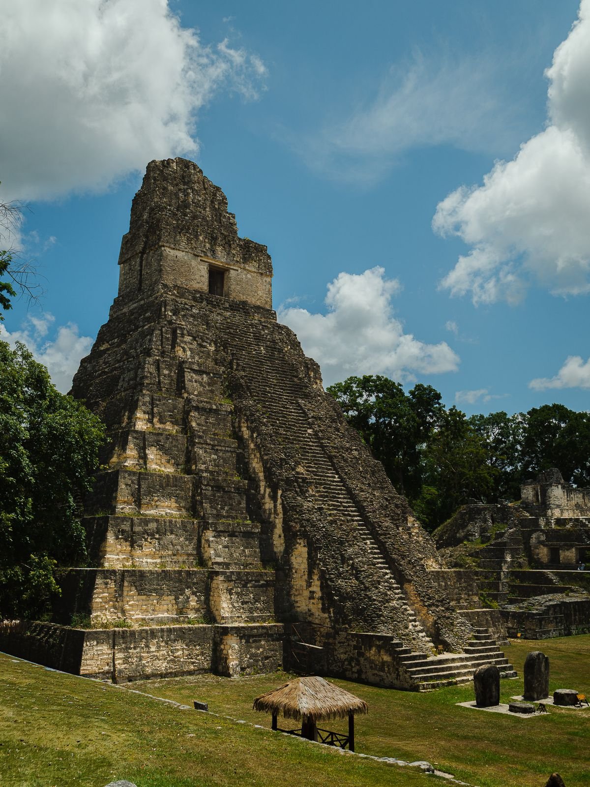 A tall stone Maya temple with a steep central staircase rises above a green lawn under a partly cloudy sky.