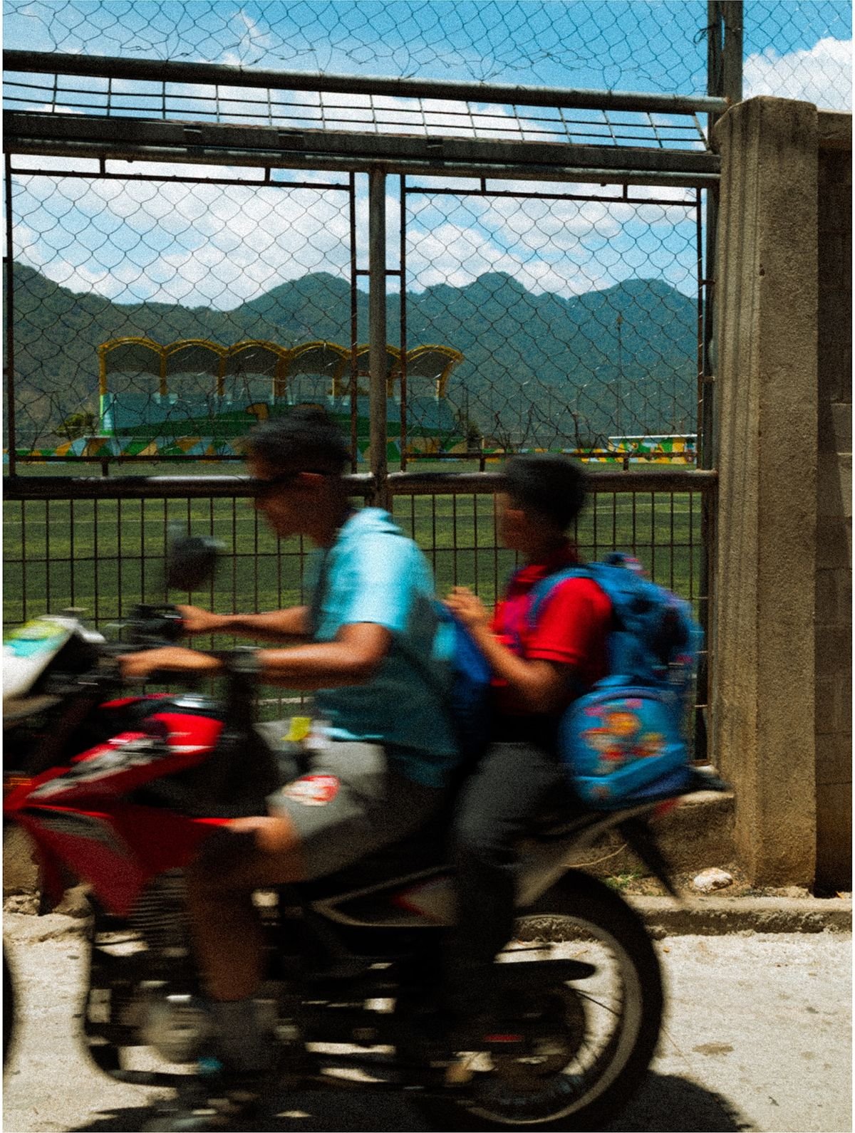 Two boys ride a red motorcycle past a fenced soccer field, blurred with motion.