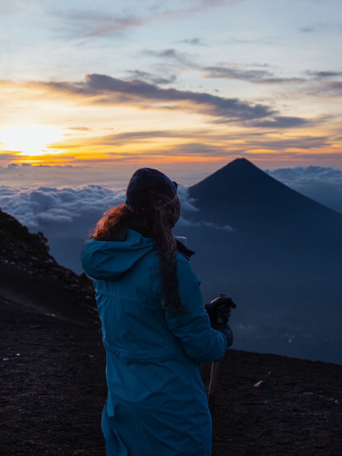 A hiker in a blue jacket stands on dark volcanic ground at sunrise, facing a cone-shaped volcano above a sea of clouds.