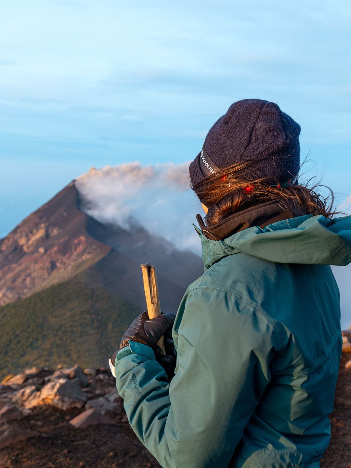 A hiker in a knit hat and green jacket looks toward a volcano as smoke drifts from its peak.
